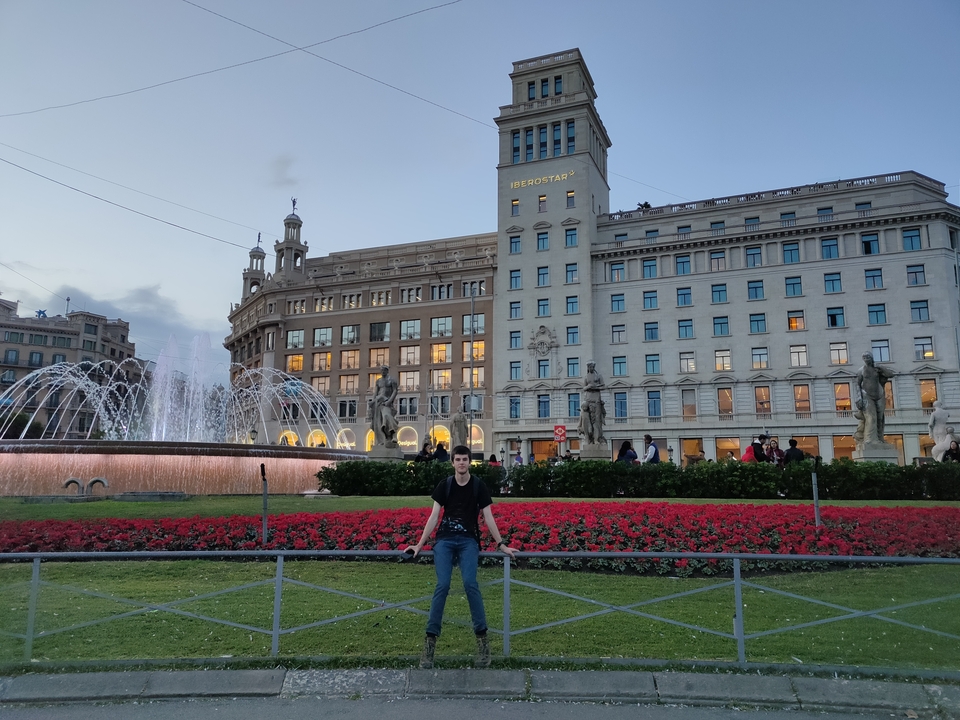 Personne posant sur une place publique avec des fleurs et une fontaine.