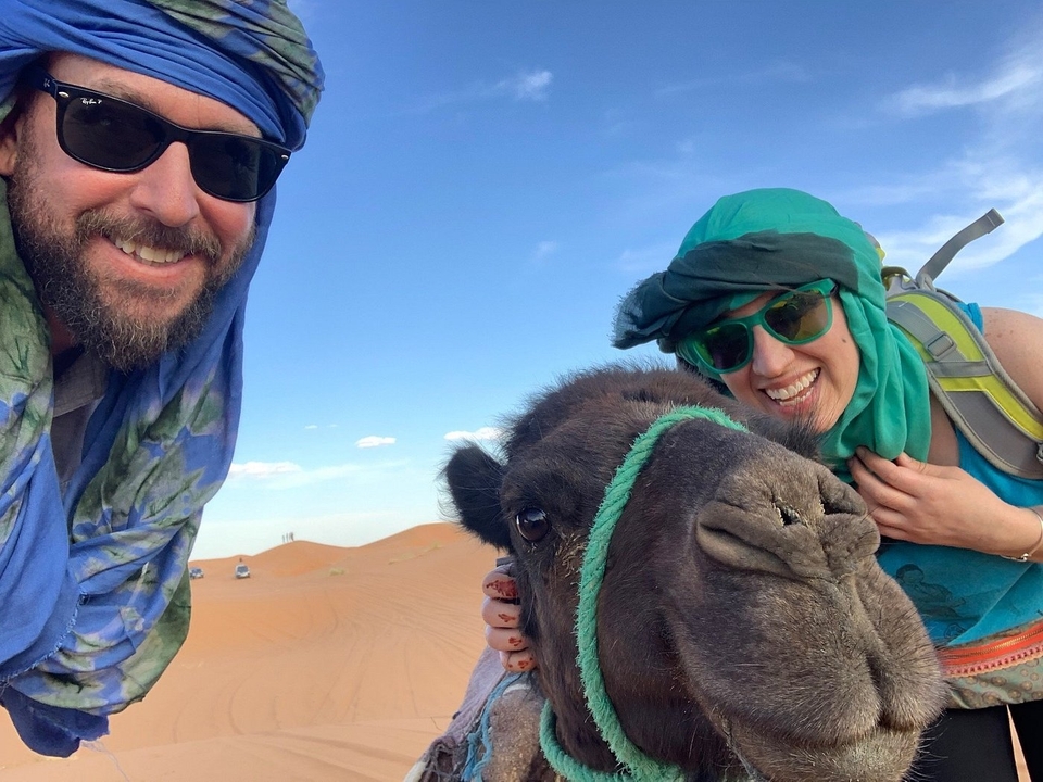 Twee glimlachende personen met een kameel in de woestijn.