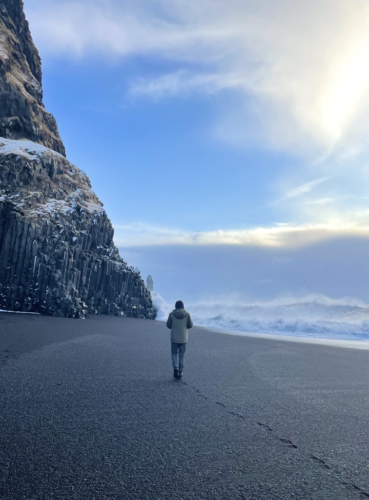 Person standing near a towering cliff at a rocky beach.