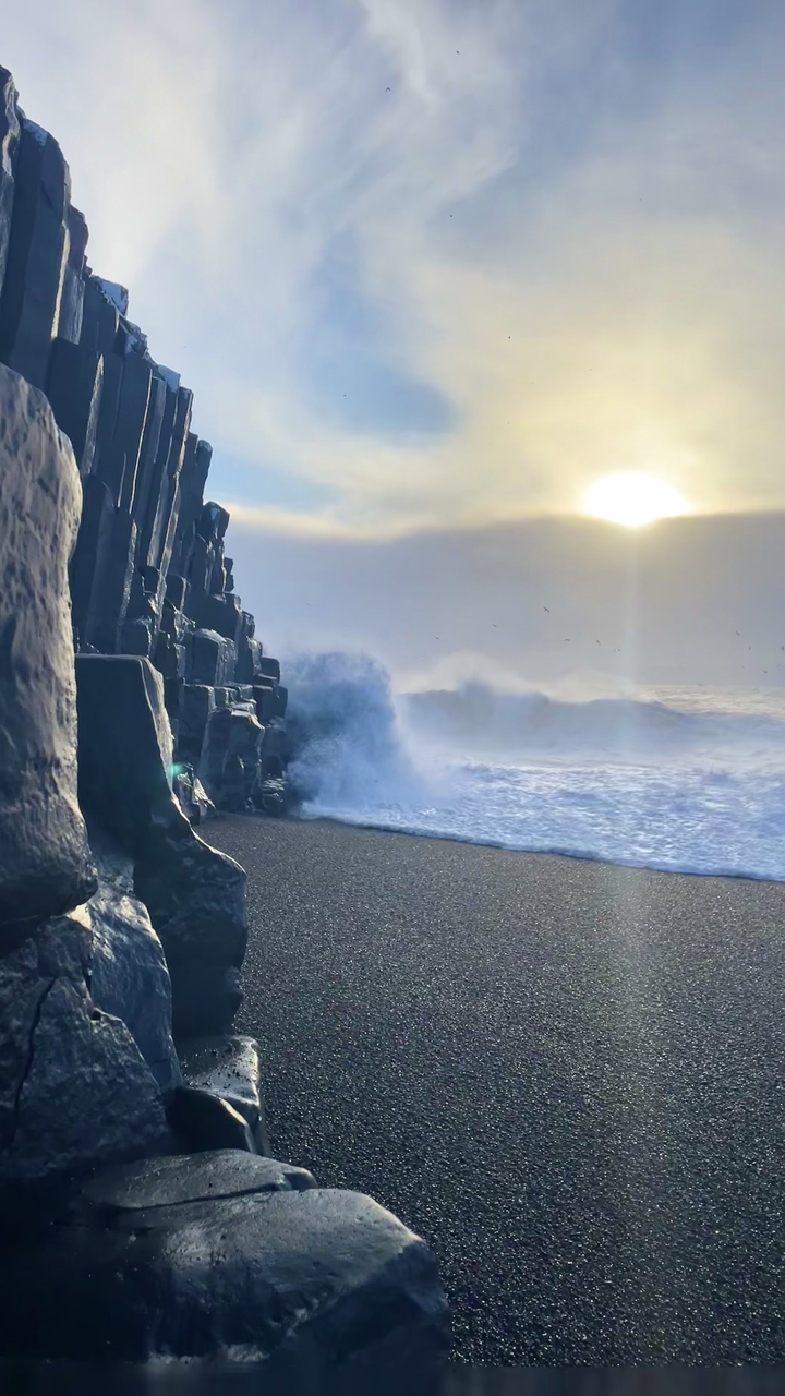 Waves crashing onto a rocky shore with sunlight breaking through clouds.