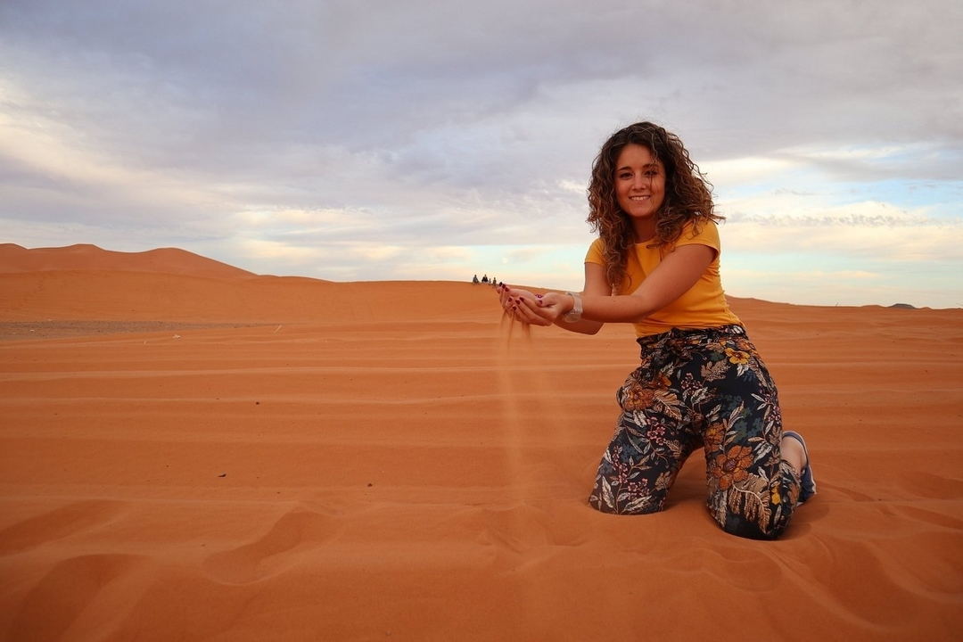 A woman kneeling in the sand dunes holding sand with a soft sky above.