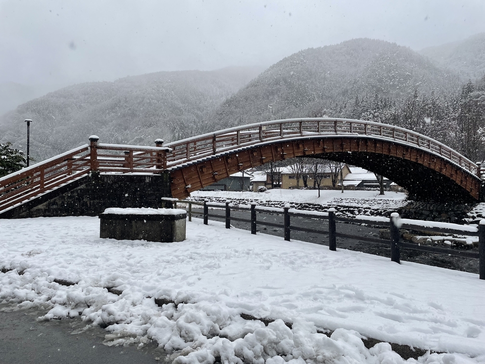 Snow-covered bridge over a river with mountains in the background.