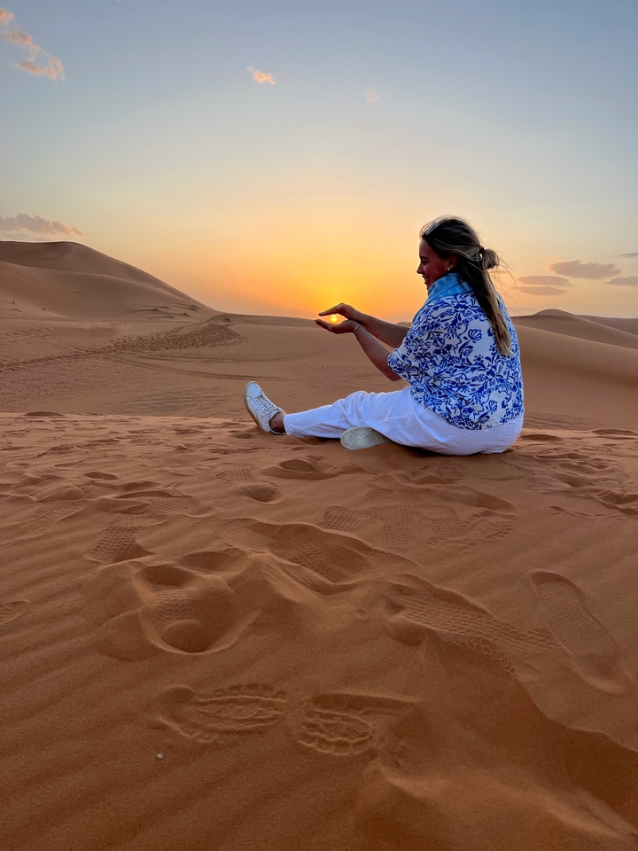 Woman sitting on sand dunes at sunset, playfully holding the sun.