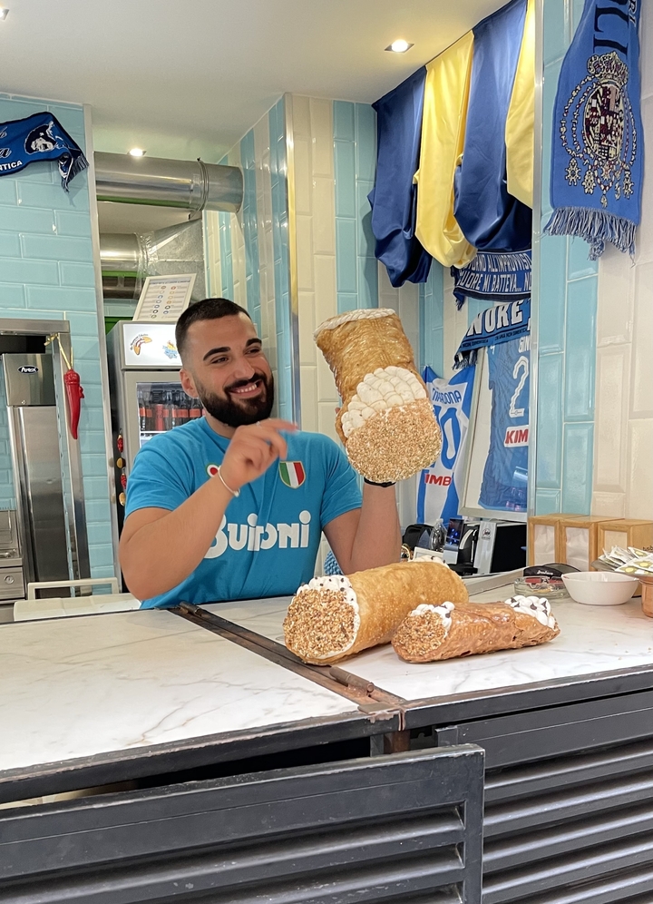 Man holding large cannoli in a cafe setting.