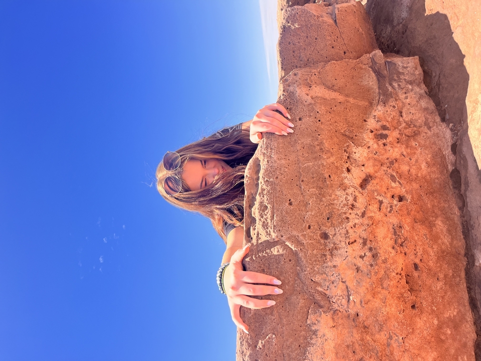 Person leaning over a cliff edge with a clear blue sky.