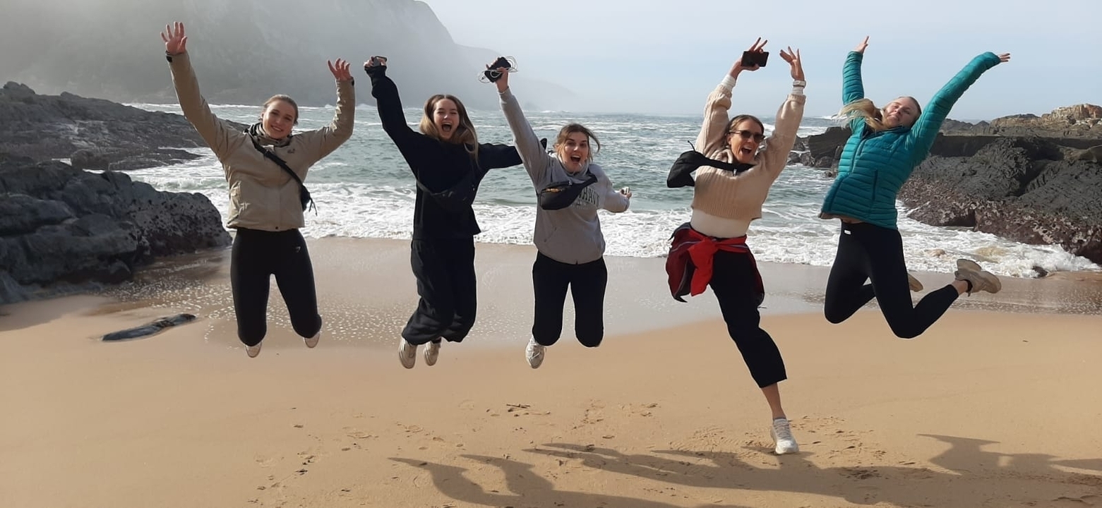 Group of people jumping on a beach with a rocky coastline.