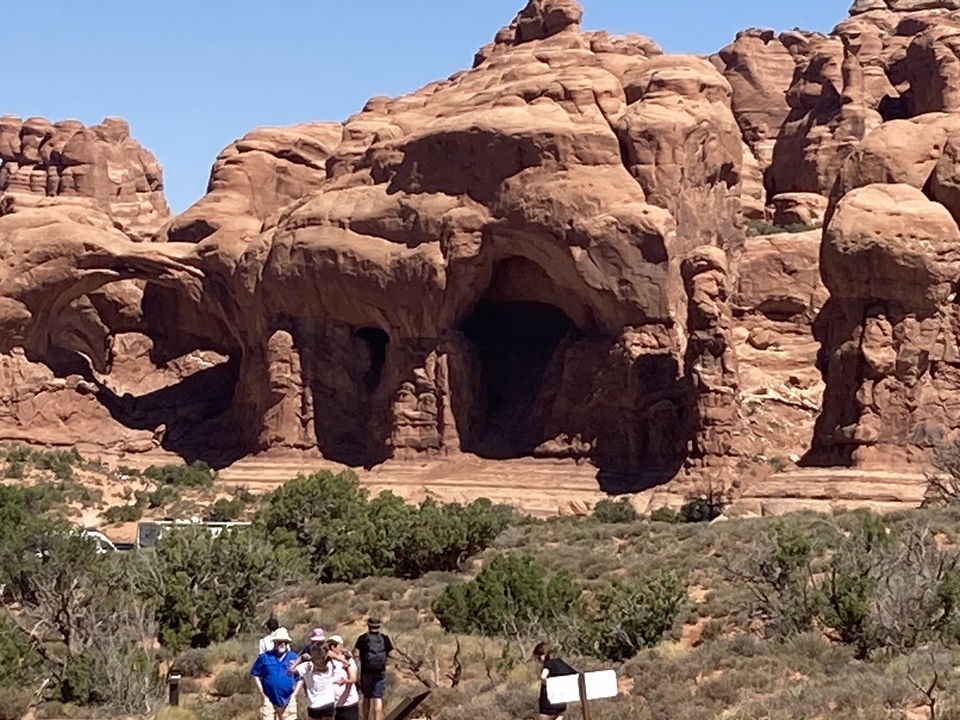 Natural rock arch in a desert setting with bushes and blue sky.