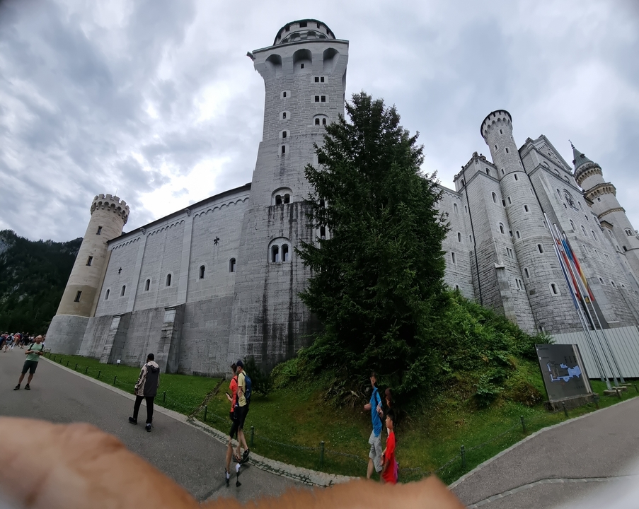 Un grand château de pierre avec des tours et des drapeaux. Des gens sont visibles le long du chemin.