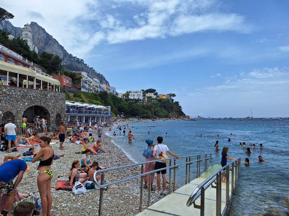 Crowded beach with tourists and boats by a rocky shore.