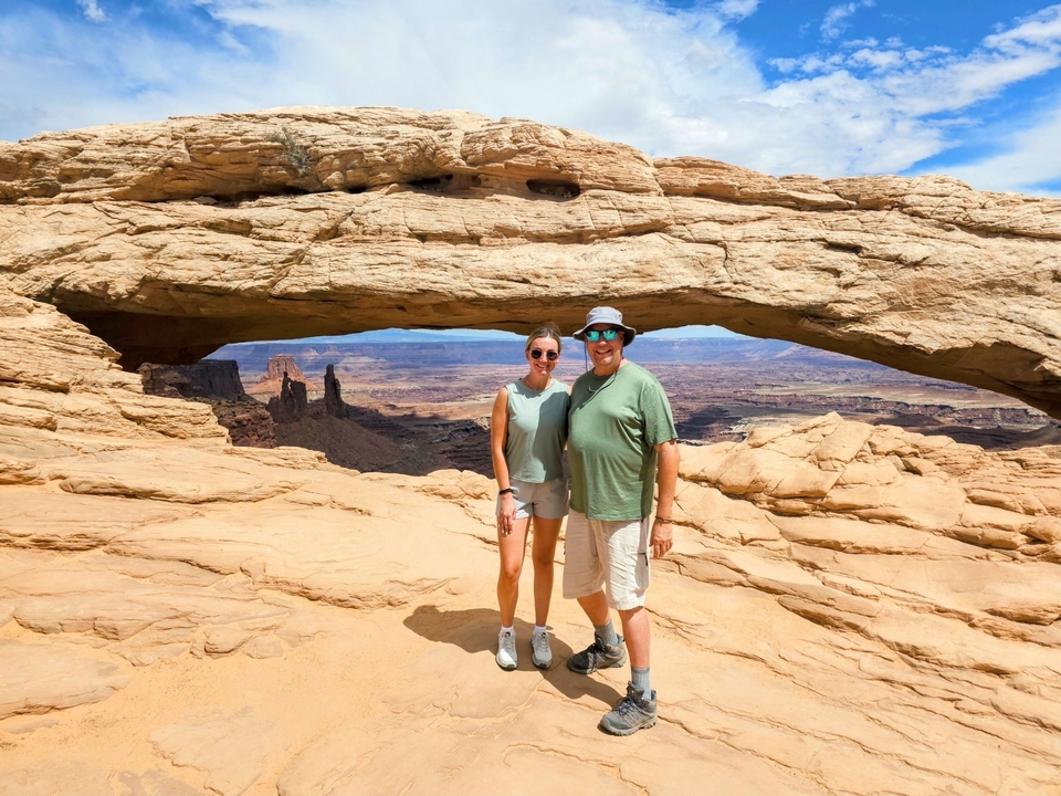 Un couple debout sous une arche rocheuse naturelle avec un vaste paysage derrière.