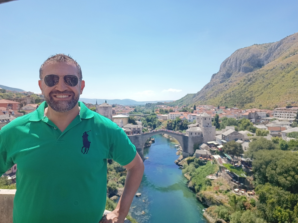 A man smiling at a historic bridge over a river.