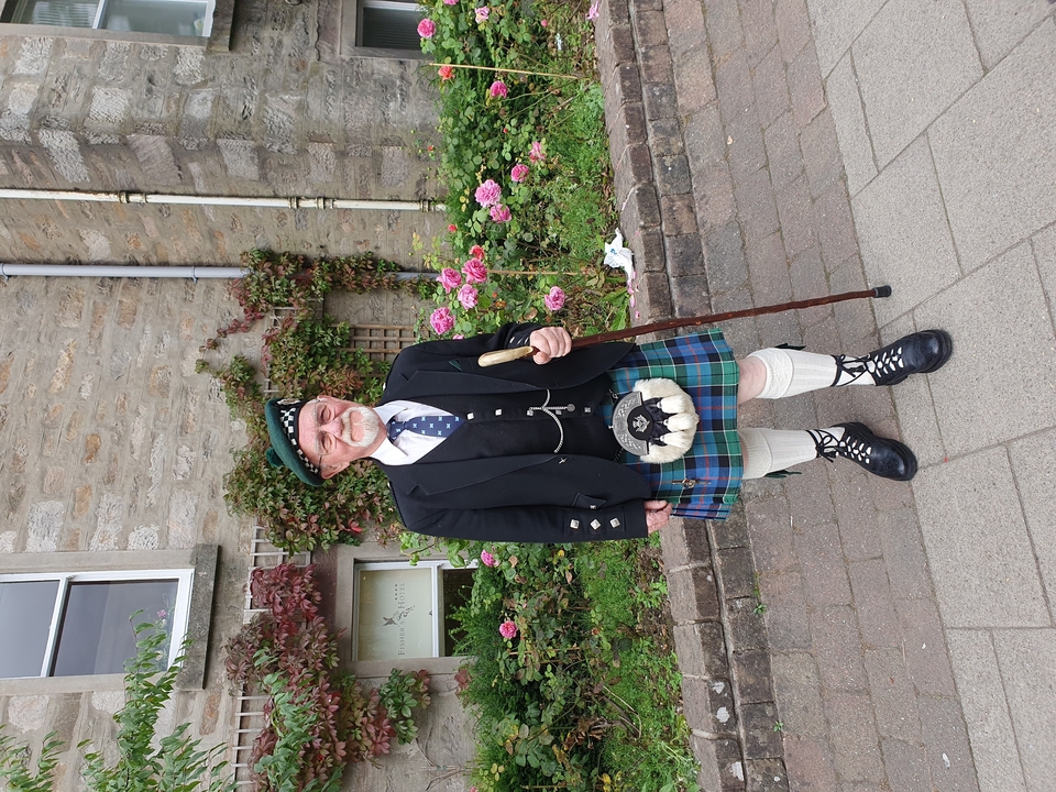 Man in traditional Scottish attire holding a cane, standing outdoors.