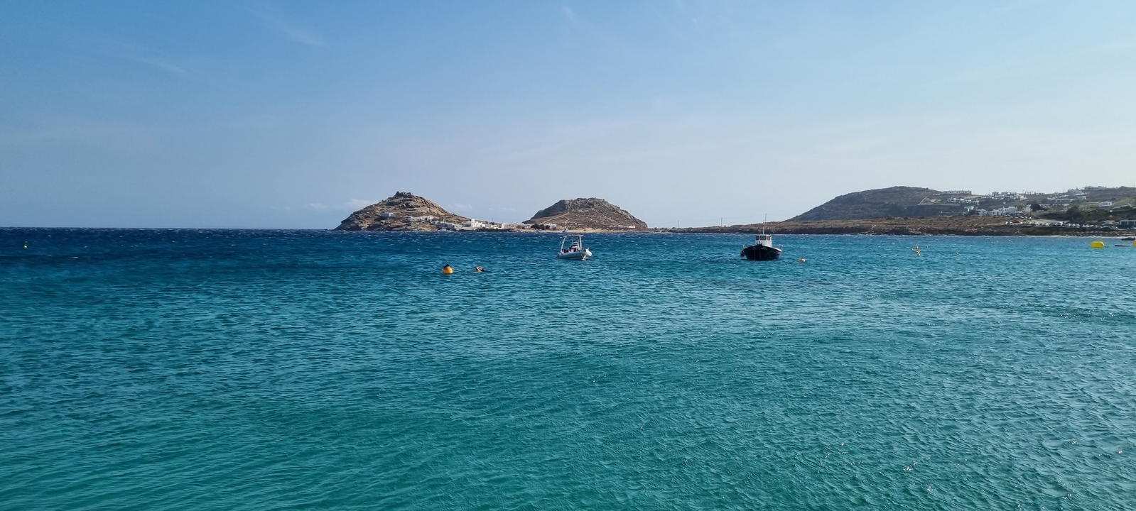Clear blue sea with small boats and distant hills.