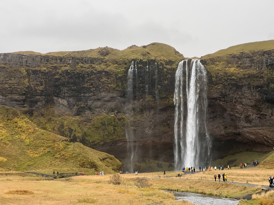 Tall waterfall with people at the bottom, surrounded by green cliffs.