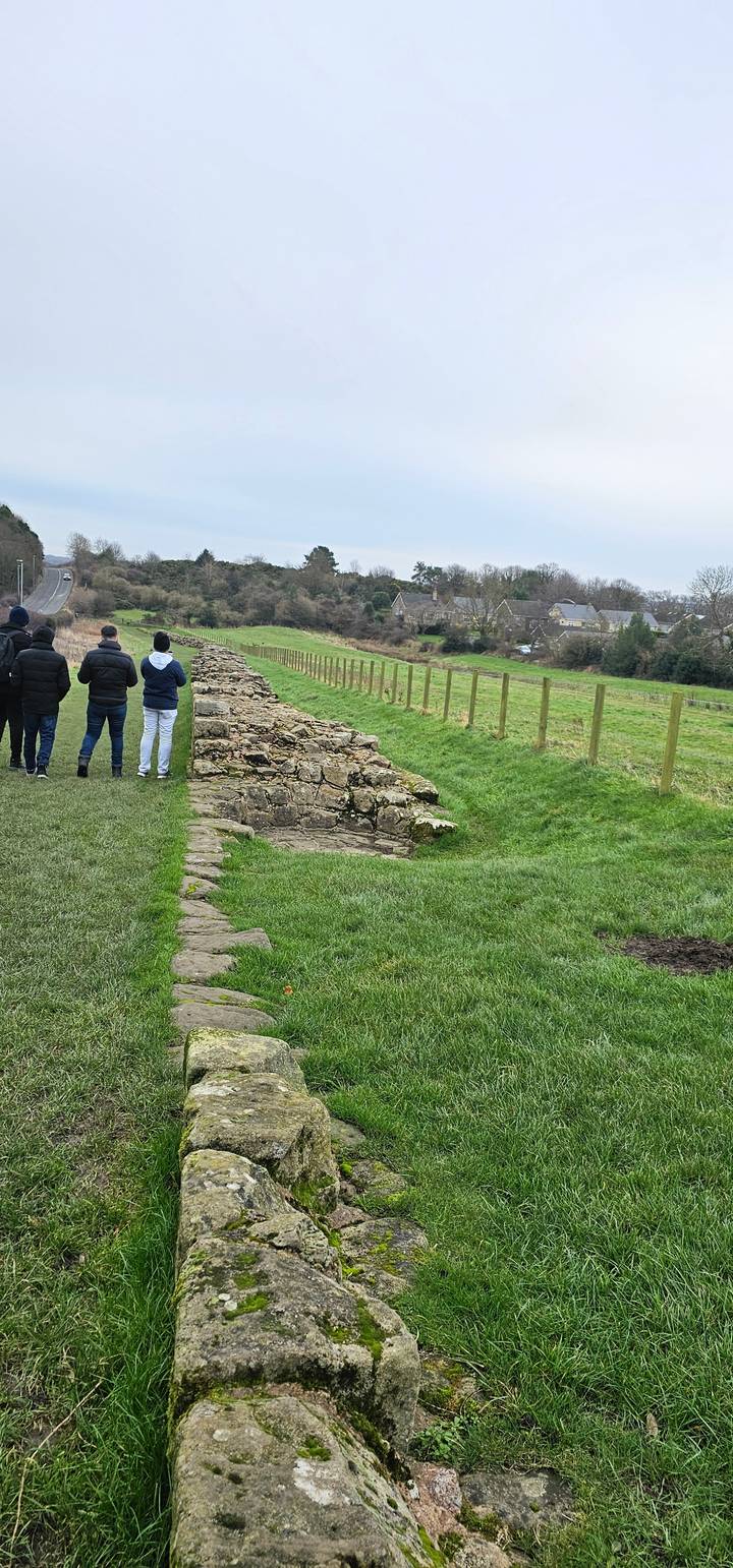 People walking along a stone wall with green fields.