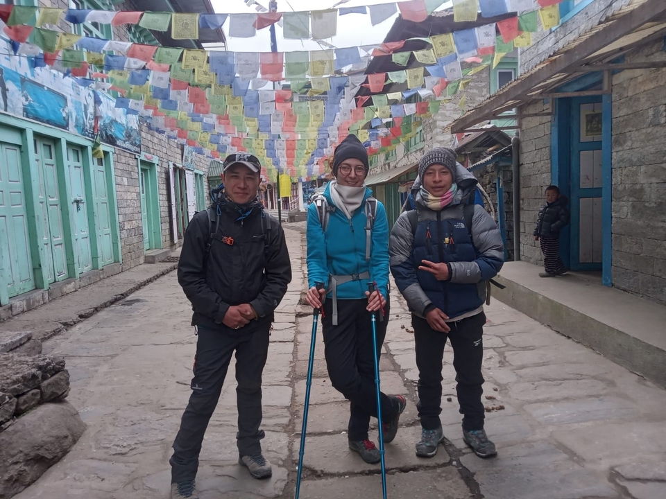 Three trekkers posing in a Nepali town with colorful flags above.