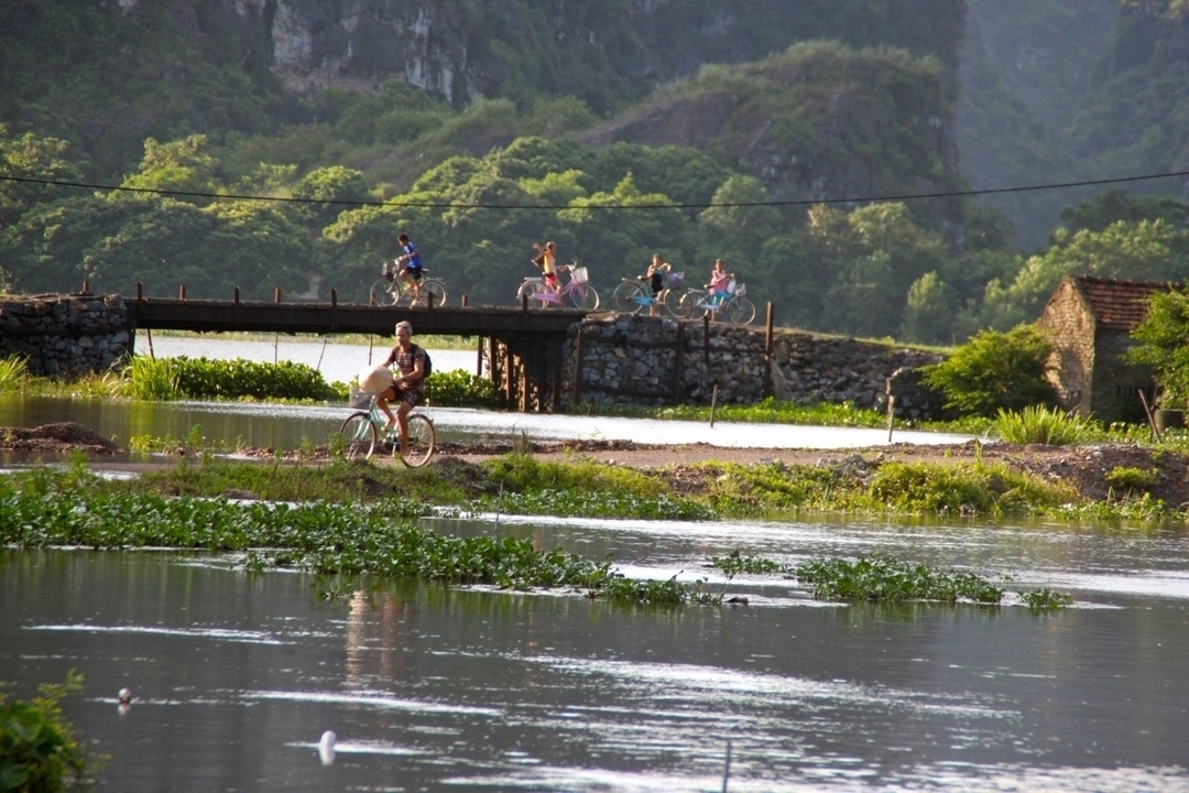 People cycling along a path with a river and mountains.