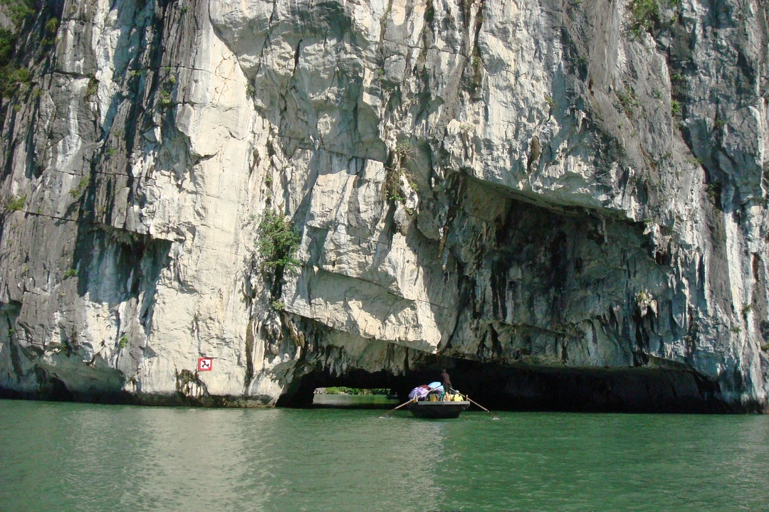 Small rowboat entering a dramatic limestone arch along emerald waters and sheer karst walls