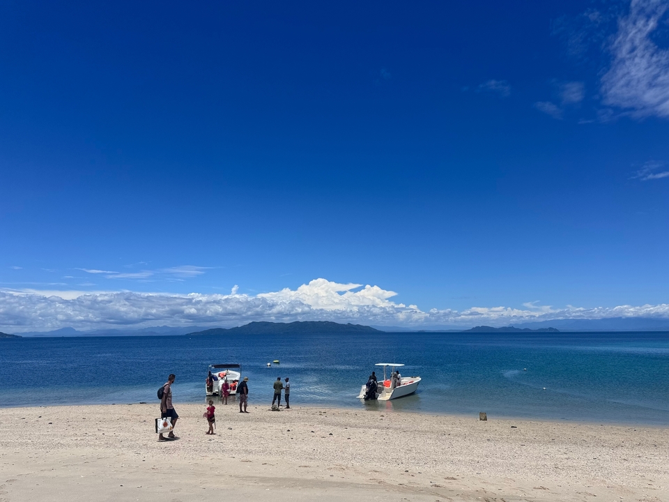 People on a beach with boats on a clear blue sea and sky.