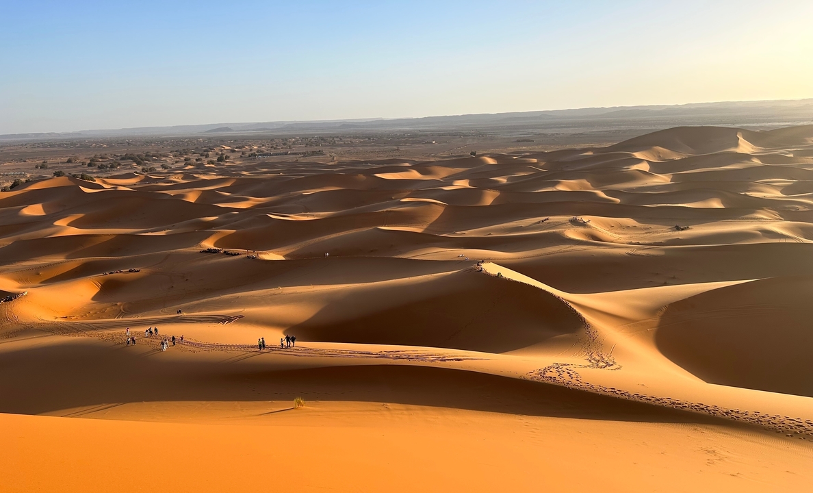 Vue expansive de dunes de sable au coucher du soleil avec de minuscules silhouettes visibles.