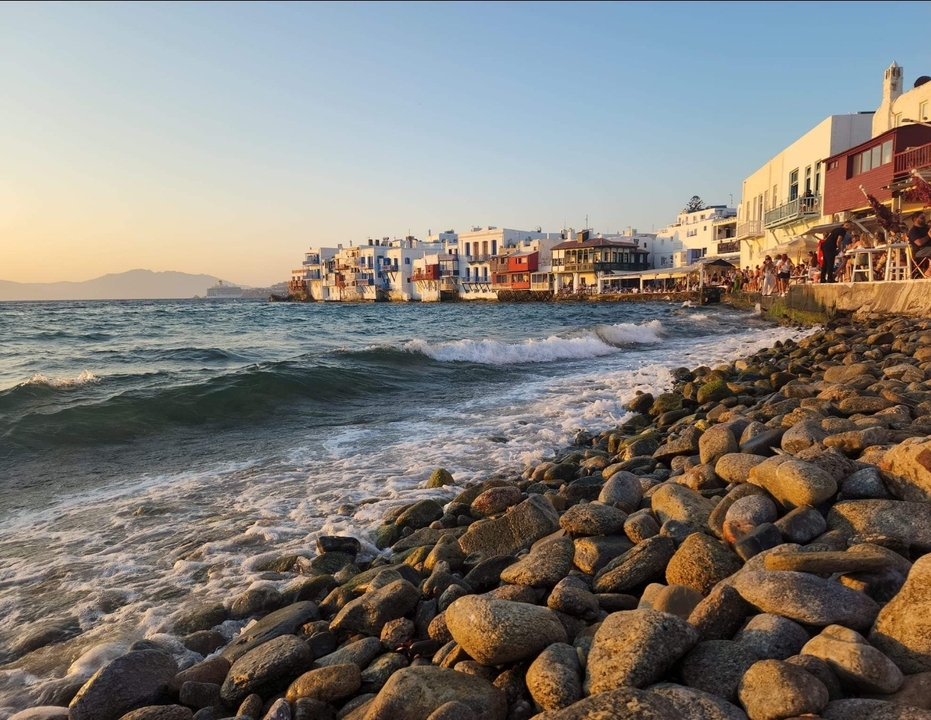 Coastal town with white buildings by the sea and rocky shore under a warm sky.