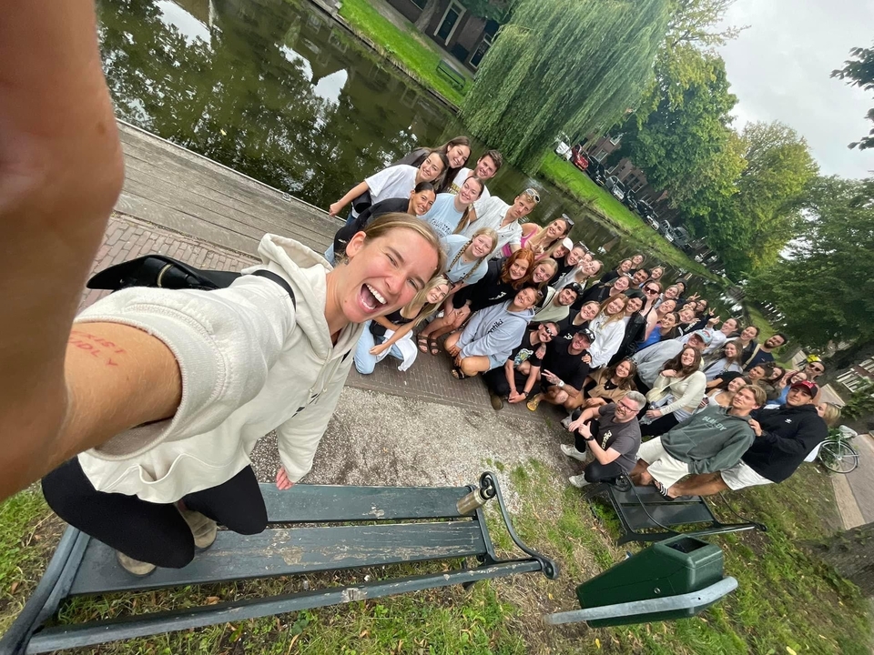 Large group taking a selfie in a park with trees and water.