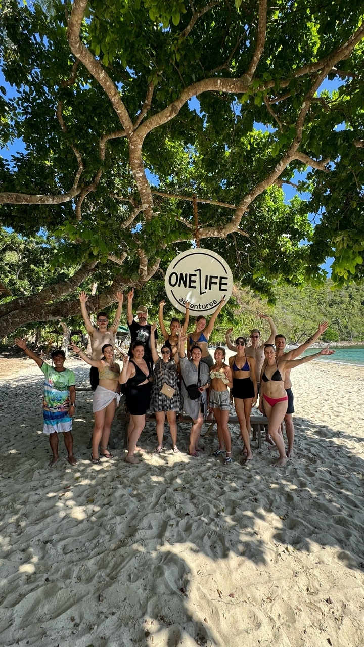 Groupe de personnes posant sous un panneau 'One Life Adventures' sur une plage.