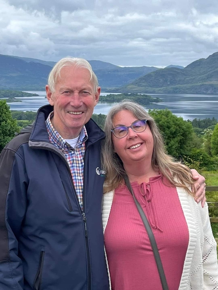 A couple posing in front of a lake with greenery in the background.