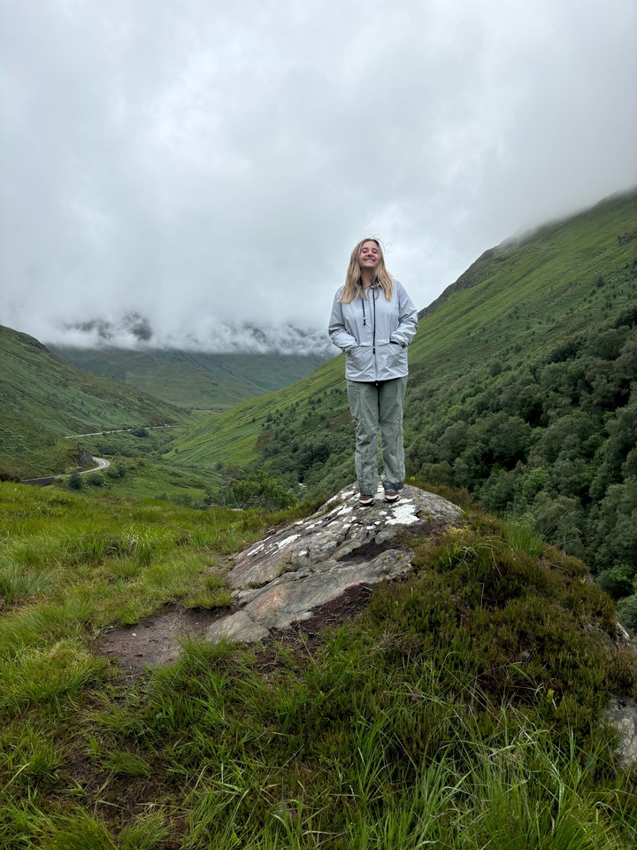 Personne debout sur un rocher avec des collines vertes et des nuages brumeux en arrière-plan.