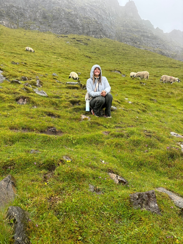 Personne accroupie sur un terrain herbeux avec des moutons qui broutent à proximité.