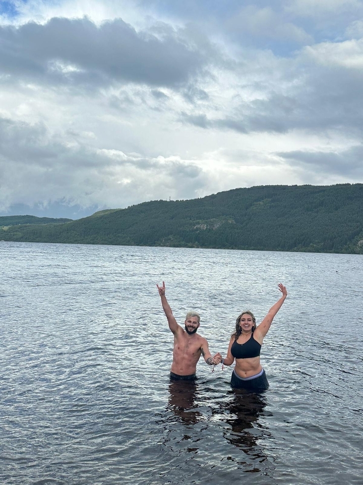 Two people swimming in a lake with forested hills.