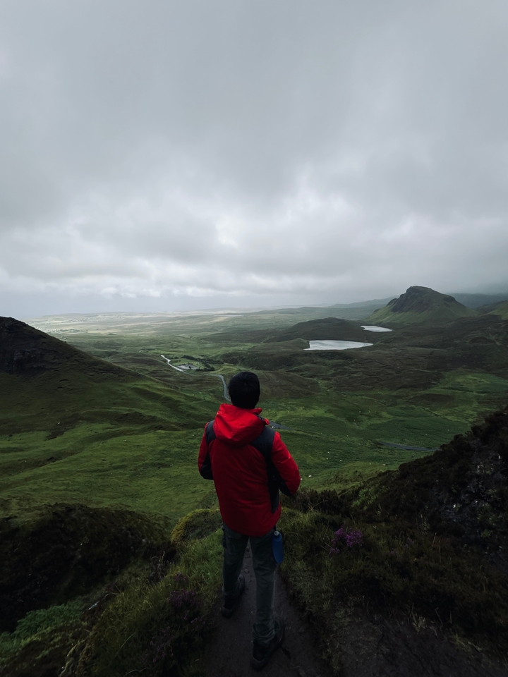 Person in a red jacket overlooking a vast green landscape with small lakes.