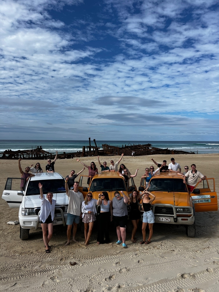 Group of people posing with cars on a beach by shipwreck.