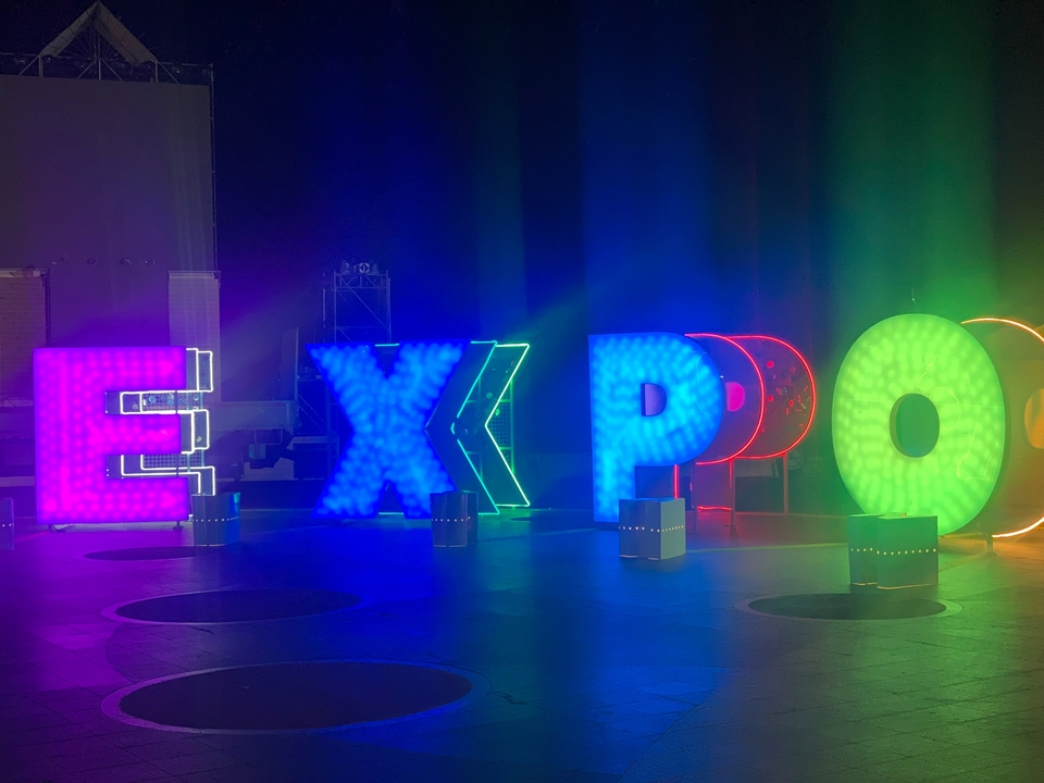 Lettres colorées lumineuses formant le mot 'EXPO' dans un espace sombre.
