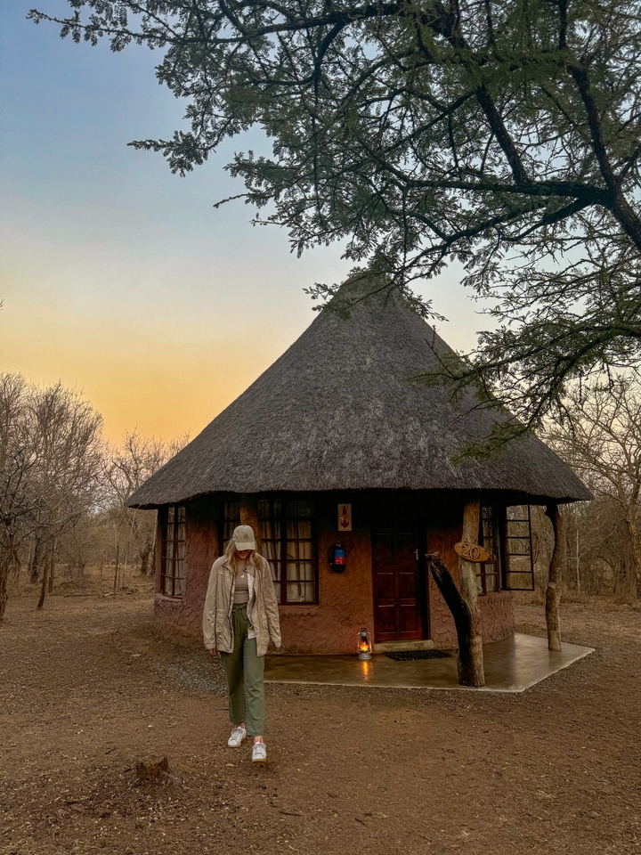 A person standing in front of a thatched-roof hut in the savannah.