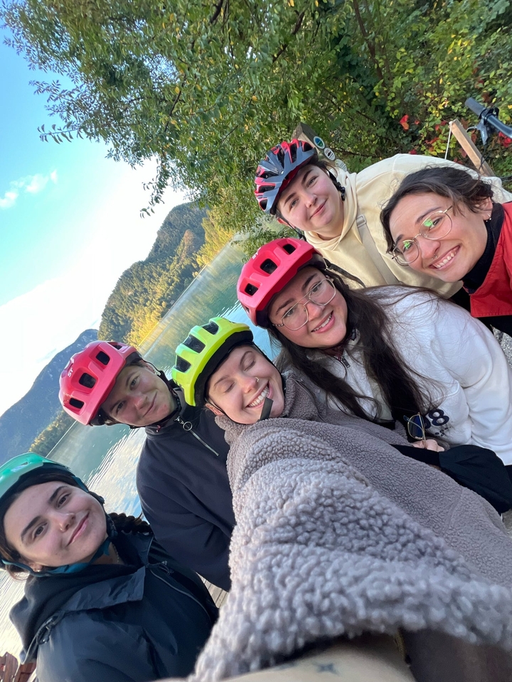 Group selfie of friends wearing helmets, by a lake.
