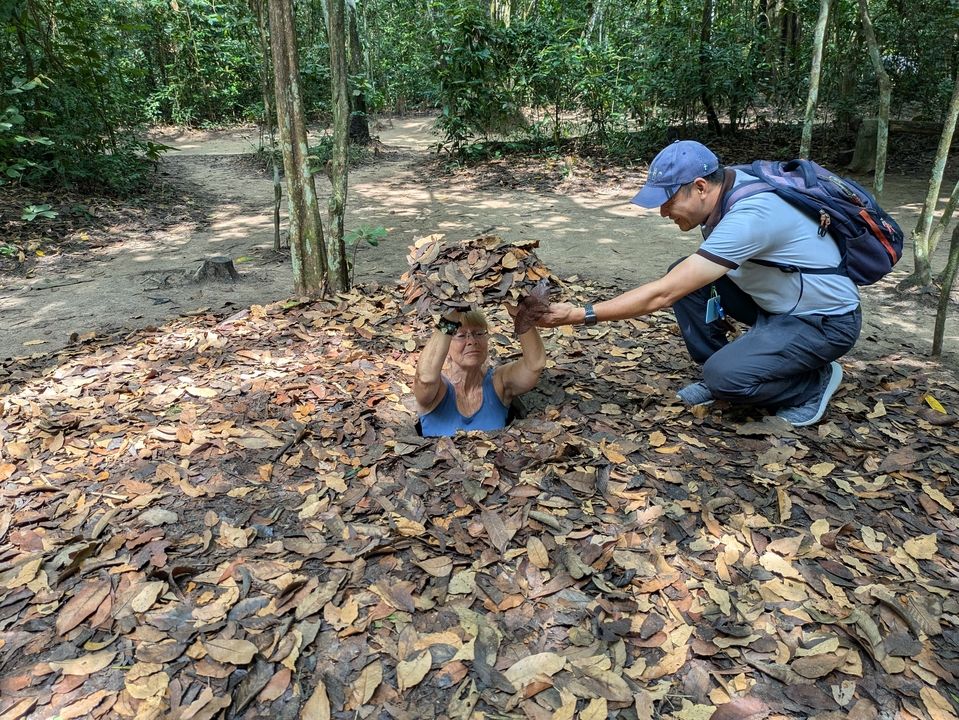 Person fitting into a small hole covered with leaves, assisted by another person.