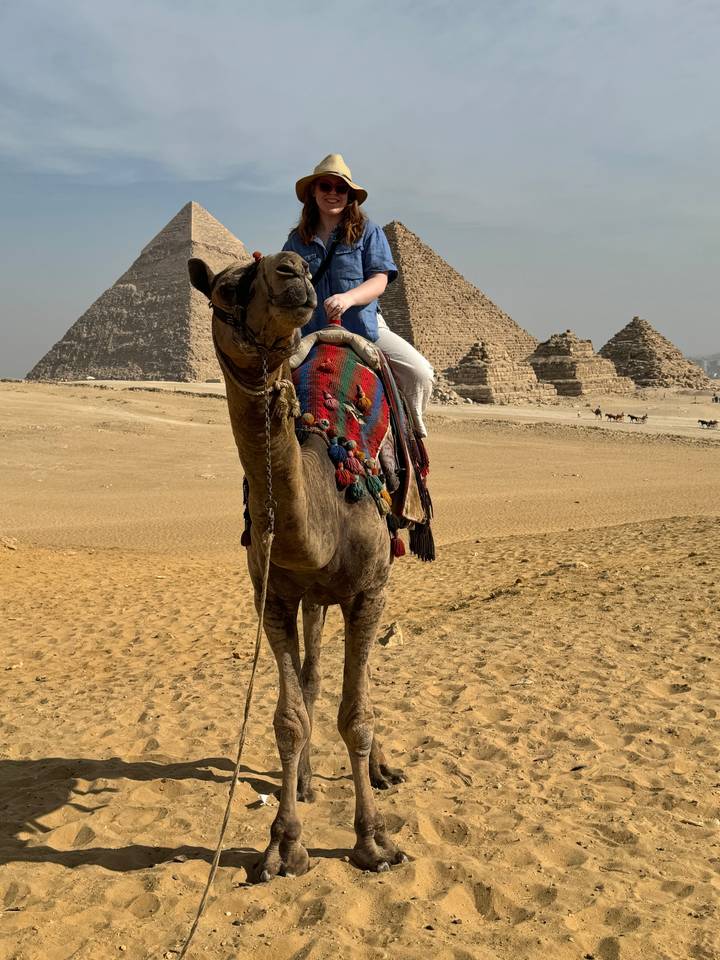 Person riding a camel with pyramids in the background.