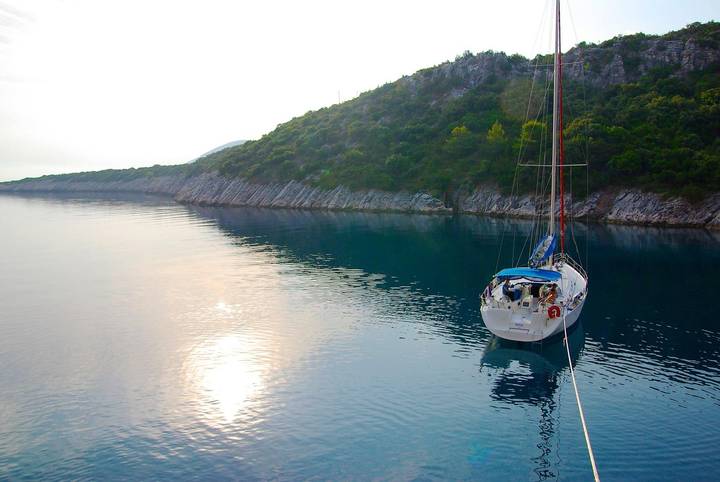 Sailboat anchored in a calm bay at sunset, with a rocky shoreline and tranquil water.
