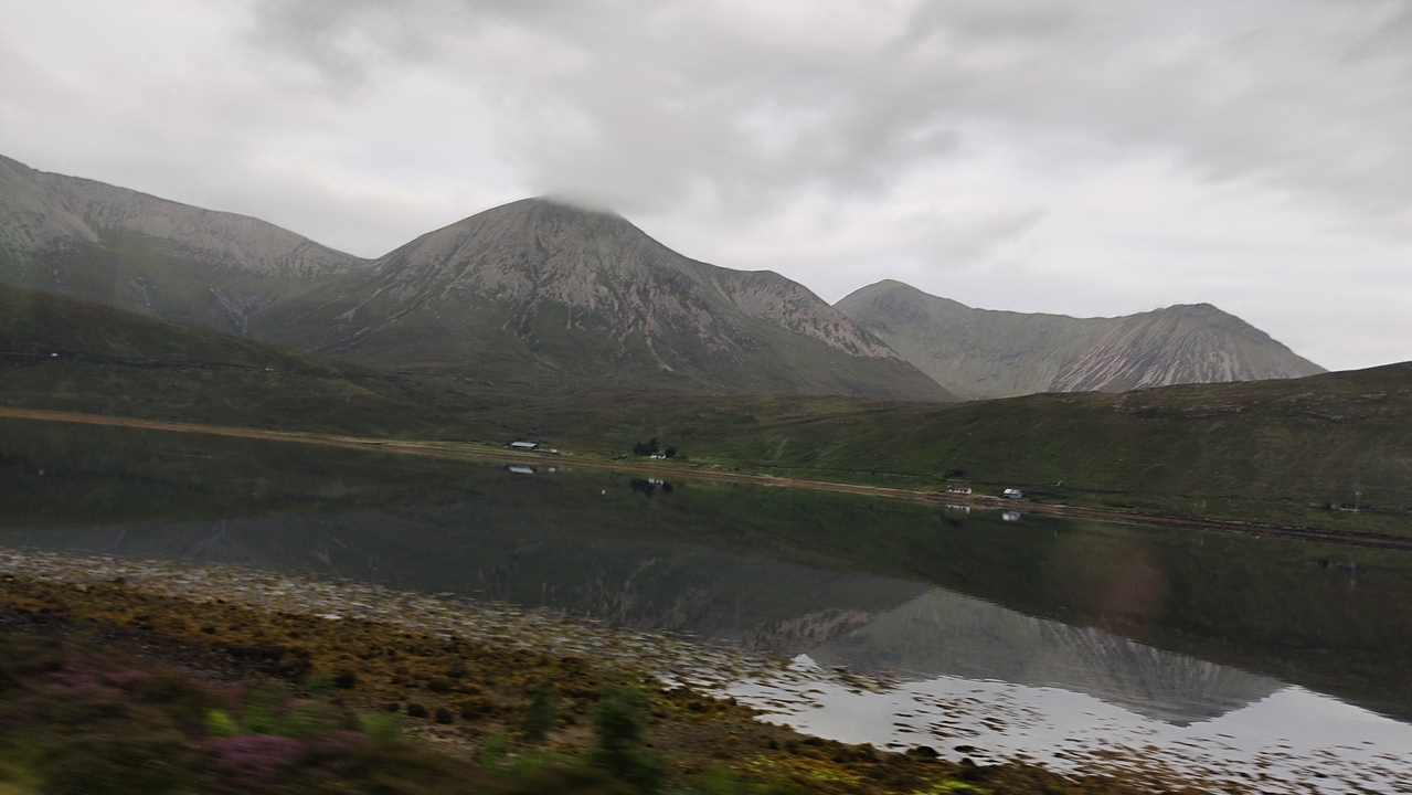 Mountain range reflected on calm water under overcast skies.