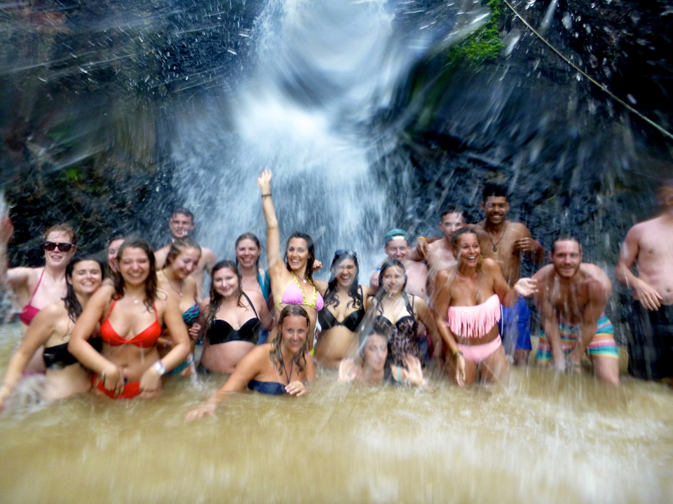 Large group cheering beneath a tropical waterfall spray in swimwear.