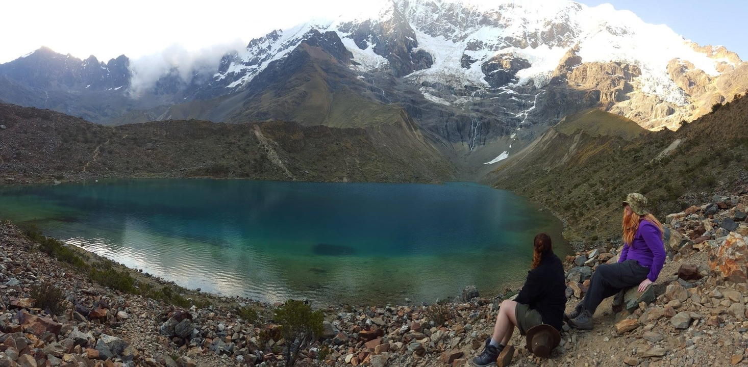 Two people sitting by a lake with snowy mountains.