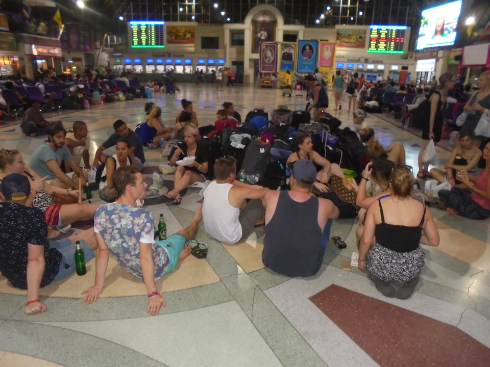 Large group of people seated indoors with luggage.