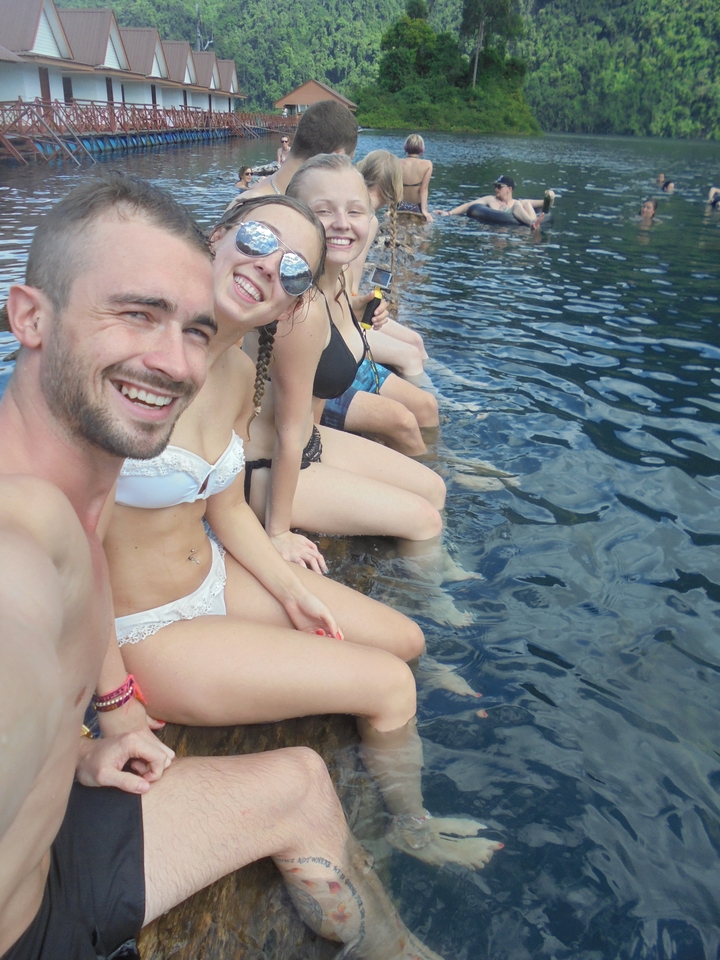 Close-up selfie of friends sitting on a wooden edge with clear blue lake water beside them.