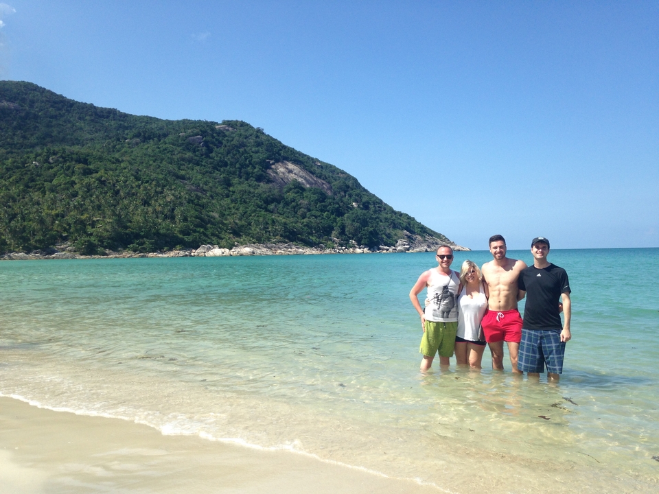Four people posing in shallow beach water with a green hill backdrop.