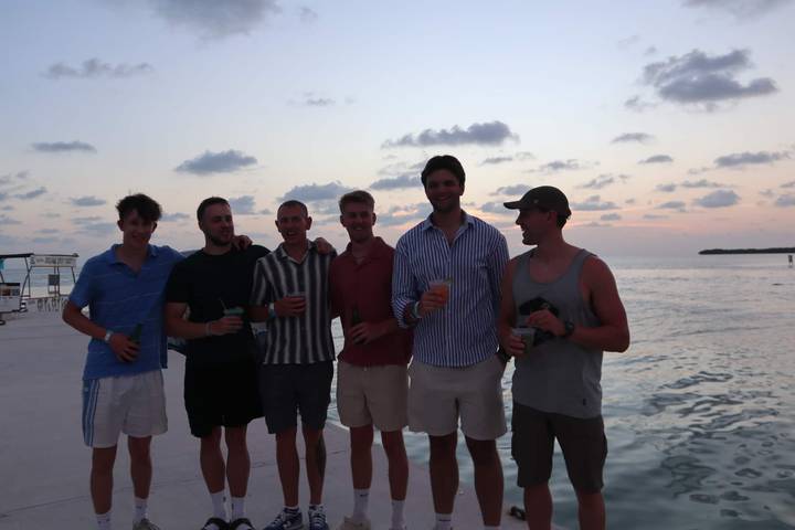 Six hommes debout près de la mer au coucher du soleil tenant des boissons.