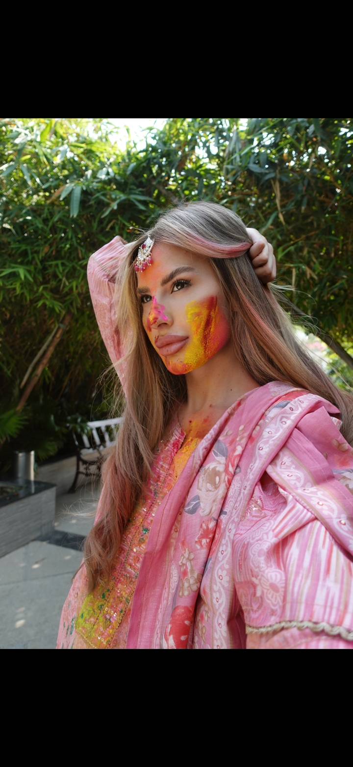A person with colorful powder on their face, likely during a traditional Indian festival, wearing a pink outfit.