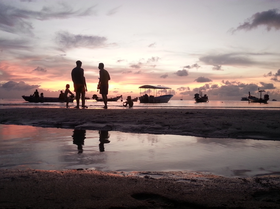 People on a beach during sunset with several boats on water.