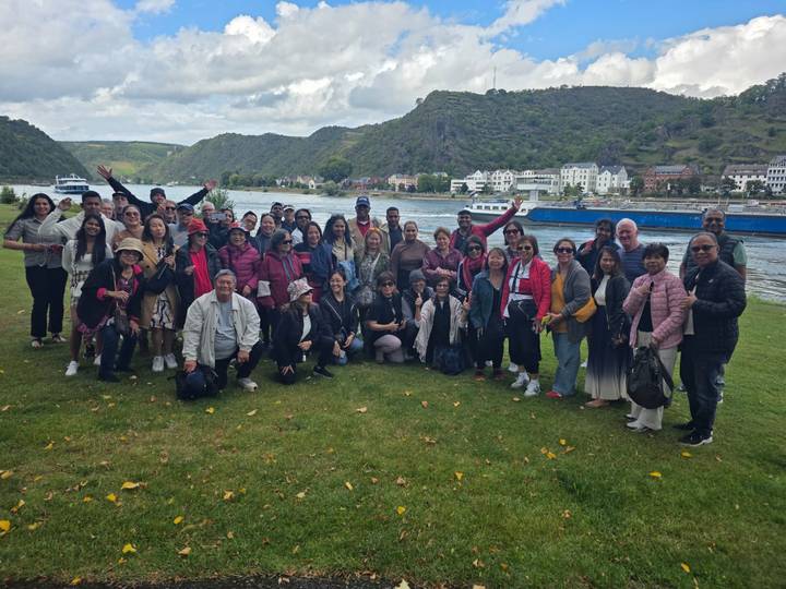 A large group of tourists posing by a river with boats and scenic hills.