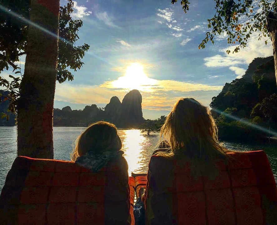 Two people enjoying sunset by the water with rocky hills.